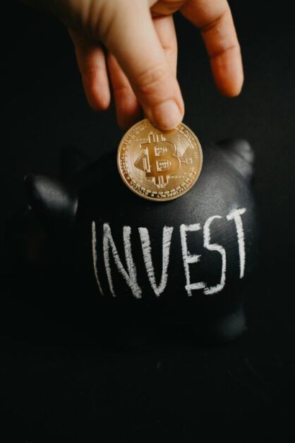 A hand holding a Bitcoin over a black piggy bank with the word “INVEST” written on it.