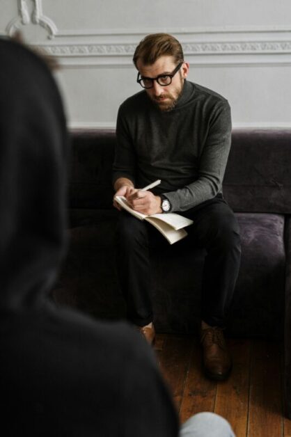 A person sitting on a dark sofa, writing in a notebook with a pen, displaying focused attention.