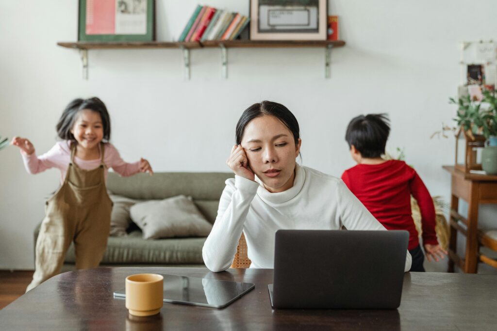 Woman working on a PC at home with two children playing in the background, feeling stressed, in a need for stress mastery