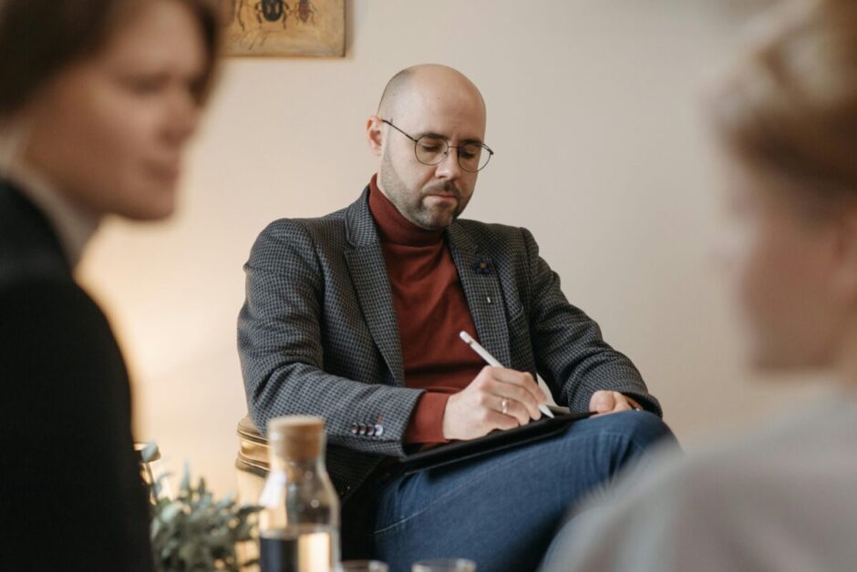 A person with a blurred face sits in a meeting, taking notes on a clipboard.