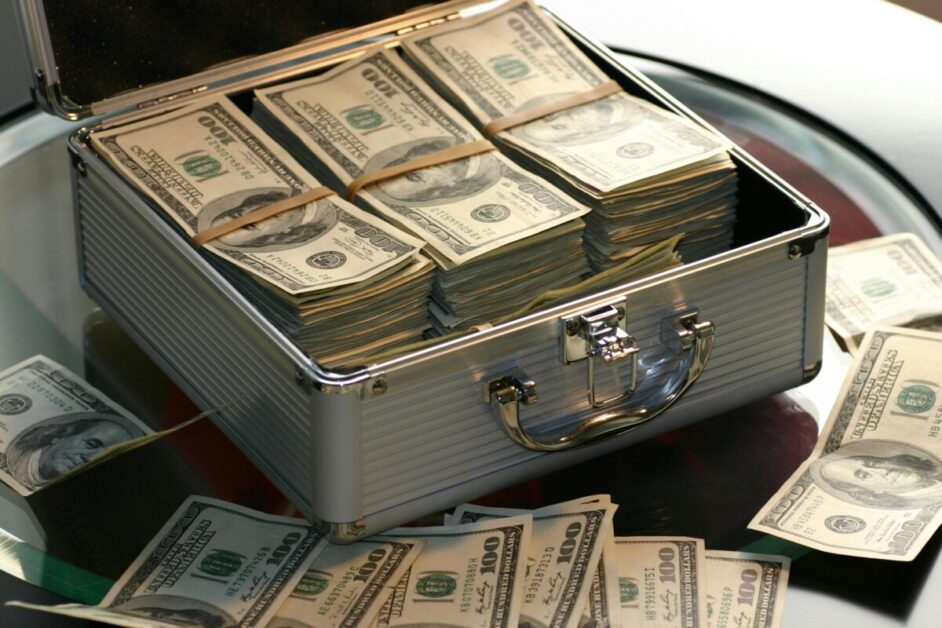 A silver briefcase filled with bundles of US dollar bills, symbolizing invest and financial growth. The open case reveals stacks of currency, with additional loose bills scattered around, indicating wealth accumulation through smart investing.