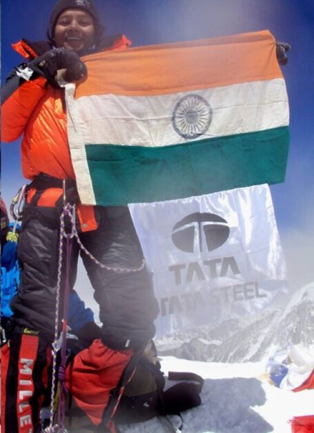 Arunima Sinha, wearing mountaineering gear and holding the Indian national flag along with a flag featuring the Tata Steel logo, standing triumphantly on a snowy mountain peak under a clear blue sky, illustrating a motivational short story.