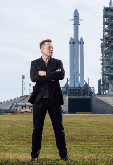 Elon Musk, dressed in a black suit, standing confidently in front of the SpaceX Falcon Heavy rocket on a launch pad, symbolizing his ambitious journey and achievements, illustrating a motivational short story.