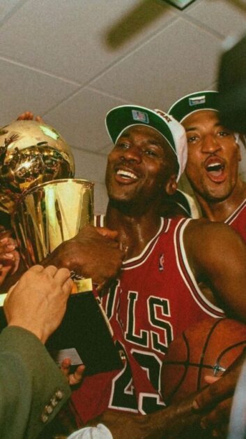 Michael Jordan, wearing his iconic red Chicago Bulls jersey number 23, holding an NBA championship trophy and a basketball, surrounded by teammates and staff in a moment of celebration, illustrating a motivational short story.