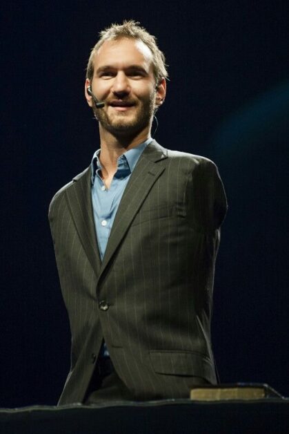 Nick Vujicic, in a suit, standing on stage with a book on the table in front of him, delivering an inspiring and motivational speech, showcasing his triumph over physical challenges, illustrating a motivational short story.