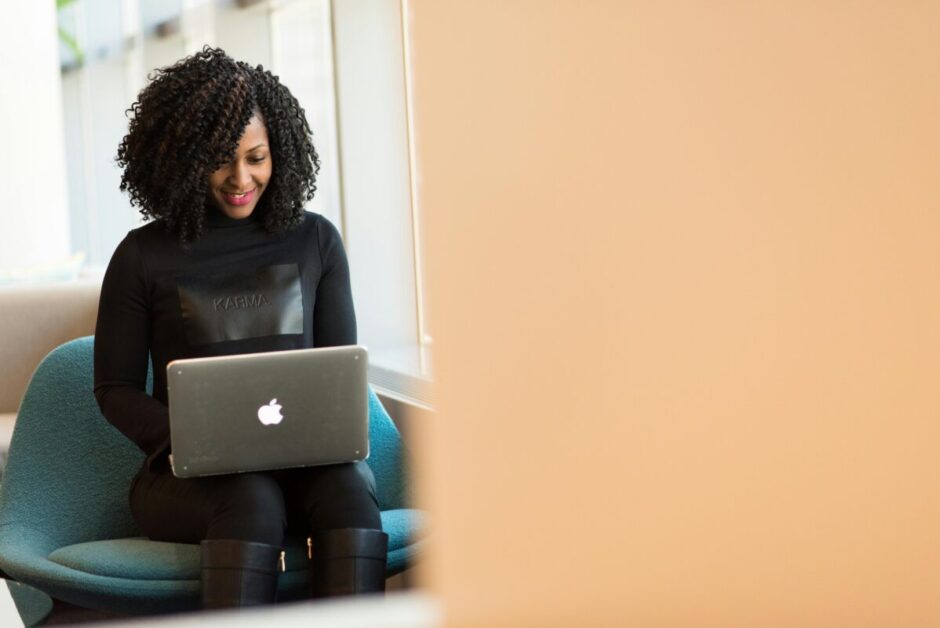 A person in professional attire sitting in a teal office chair works on a MacBook laptop with a focused smile, wearing a black sweater with 'KARMA' text. The image represents modern career development and remote work productivity in a bright, contemporary workspace setting.