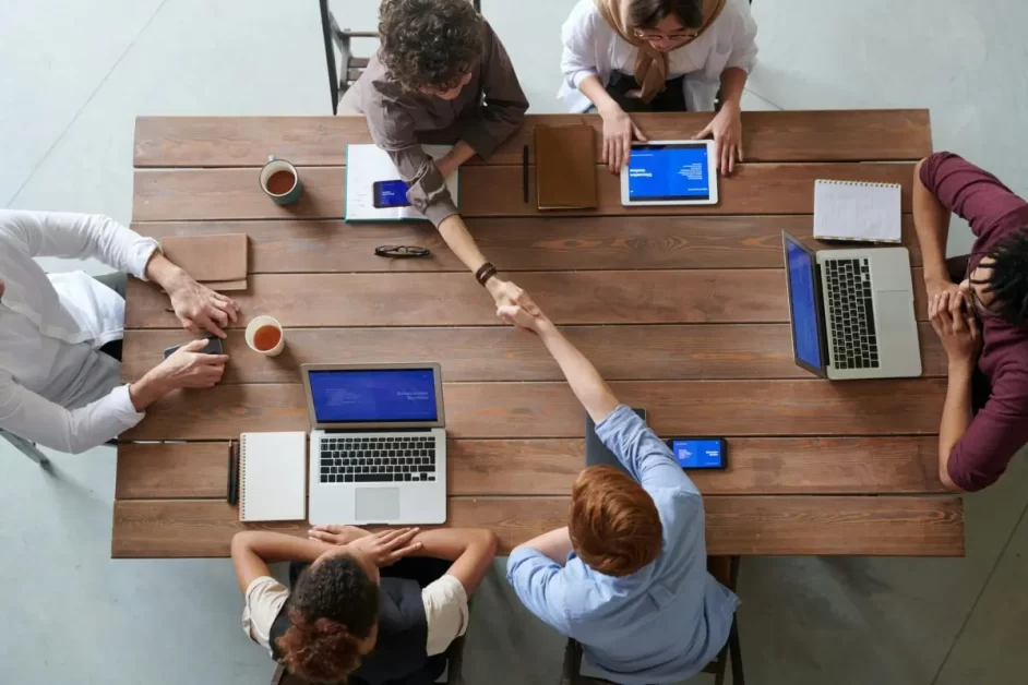 A group of professionals sitting around a wooden table, working on laptops and tablets, engaging in a meeting, with two individuals shaking hands—perfect for showcasing effective networking hacks in collaborative environments.