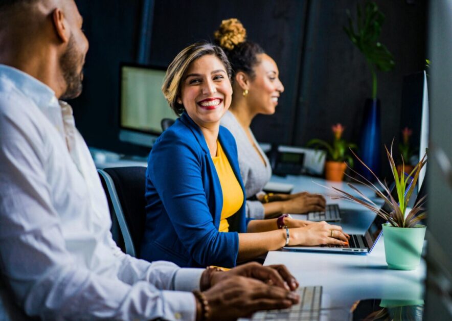 A cheerful team of professionals in an office setting, with one woman smiling while working on a laptop—ideal for demonstrating how networking hacks can foster positive workplace relationships.