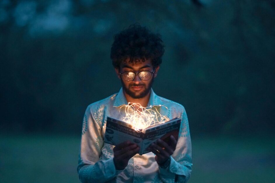 A person wearing glasses and a white patterned shirt reads a book illuminated by glowing string lights in a dark setting, symbolizing personal and career development through self-guided learning and knowledge acquisition.