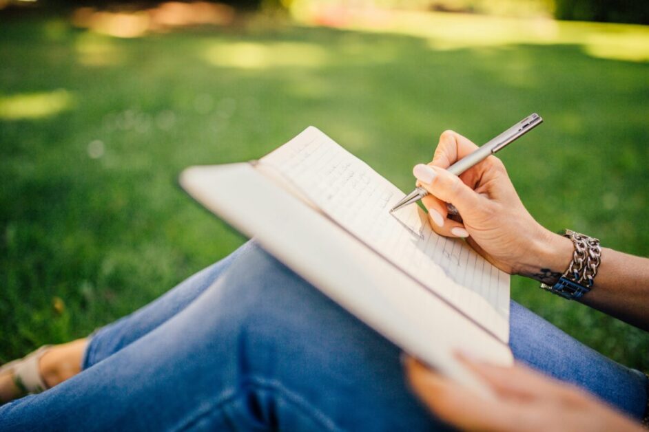 Close-up of a person sitting outdoors on a sunny day, writing in a journal, embodying the practice of positive thinking through reflection and mindfulness.